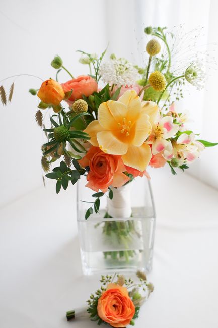 A vibrant bouquet of fresh flowers arranged in a white ceramic vase, featuring large yellow daffodils, soft peach roses, and delicate pink and white blossoms, complemented by green foliage. The arrang