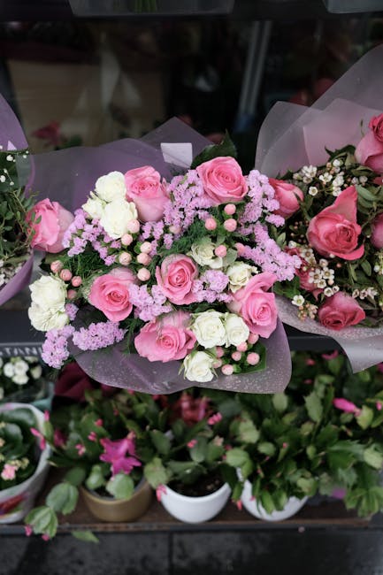 A bouquet of fresh pink roses, white carnations, and small pink and white filler flowers arranged with green foliage, wrapped in pastel pink and purple tissue paper. The arrangement is displayed on a