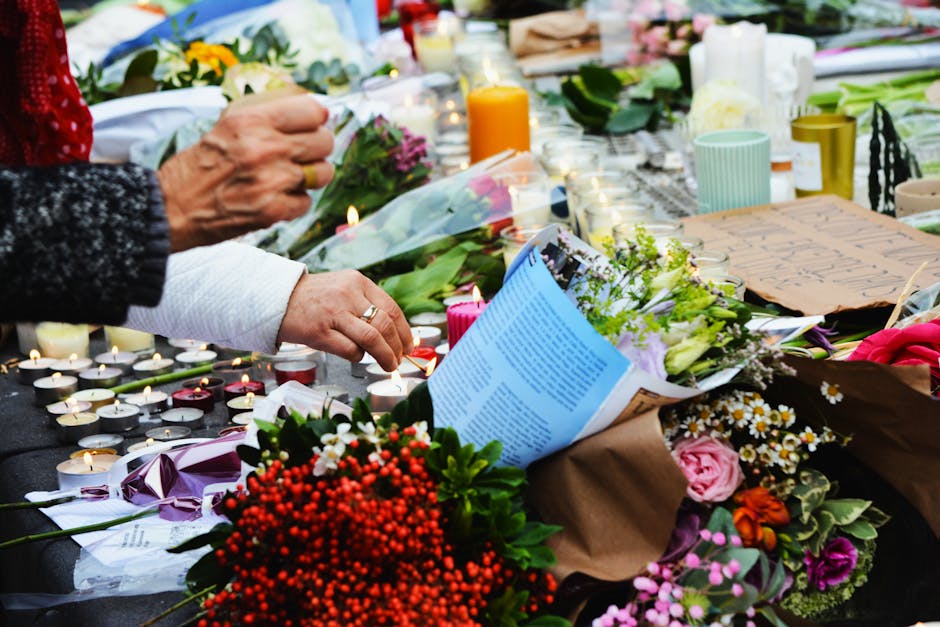 Close-up of a memorial floral tribute featuring a bouquet of vibrant mixed flowers, including pink roses, white daisies, and purple blossoms, wrapped in brown kraft paper. The arrangement is surrounde