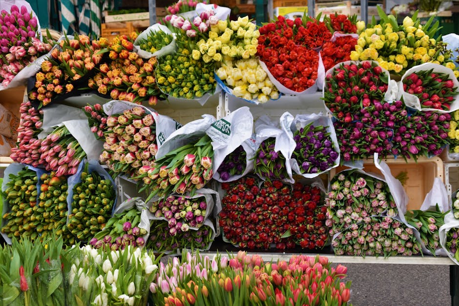 A vibrant display of fresh flowers arranged in bouquets and wrapped in white and decorative paper, featuring tulips in shades of pink, red, purple, and white, along with various other blooming flowers