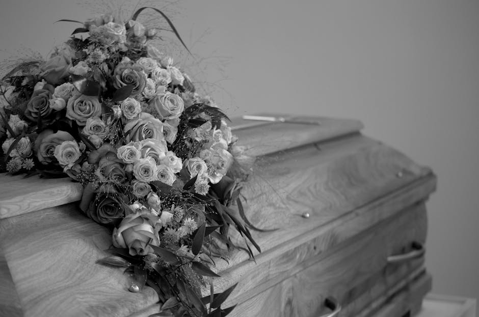 A funeral flower arrangement placed atop a wooden casket, featuring a dense bouquet of various fresh roses in shades of white, cream, and deep red. The arrangement includes greenery and delicate fille